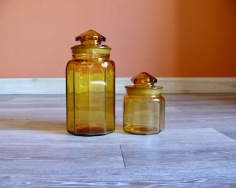 May include: Two amber glass jars with lids. The larger jar is taller and wider than the smaller jar. Both jars have a hexagonal shape.