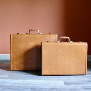 May include: Two vintage brown leather suitcases with brass hardware. The larger suitcase is in the background and the smaller suitcase is in the foreground.