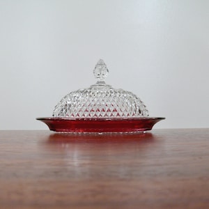 May include: A clear glass butter dish with a diamond pattern dome and a ruby red base. The lid has a decorative finial. The dish sits on a wooden surface against a white background. A classic kitchenware item.