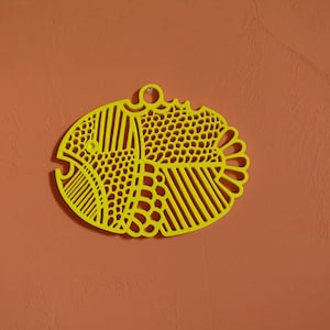May include: A yellow, fish-shaped decorative item with a detailed, openwork design. The fish has a loop at the top for hanging and is set against an orange background. The design includes various patterns and shapes.