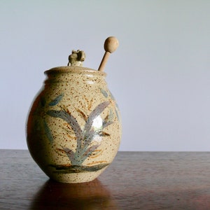 May include: A ceramic honey pot with a wooden dipper. The pot is decorated with a brown, blue, and white floral design.