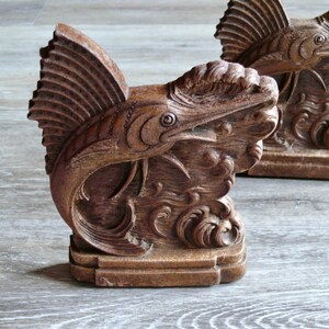 May include: A pair of wooden bookends shaped like a stylized marlin fish with intricate details. The bookends are a dark brown color and have a textured finish.