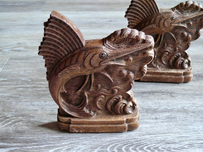 May include: A pair of wooden bookends shaped like a stylized marlin fish with intricate details. The bookends are a dark brown color and have a textured finish.