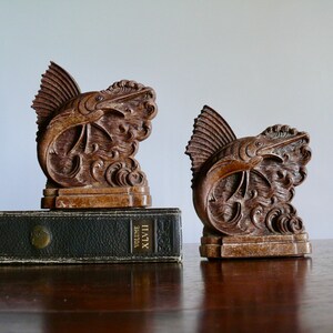 May include: A pair of wooden bookends shaped like a marlin fish leaping out of the water. The bookends are a dark brown color and have intricate details carved into the wood.