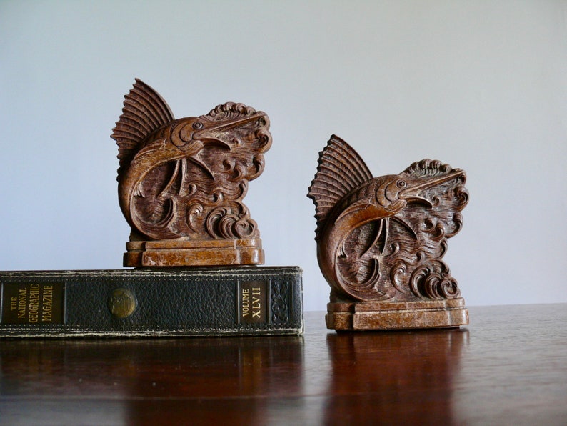 May include: A pair of wooden bookends shaped like a marlin fish leaping out of the water. The bookends are a dark brown color and have intricate details carved into the wood.