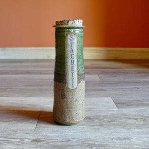 May include: A tall, ceramic spaghetti jar with a cork lid. The jar is cylindrical with a green and brown color scheme. The word "SPAGHETTI" is vertically printed on a white label in blue letters.
