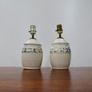 May include: Two ceramic table lamps with a cream-colored base and a white top. The lamps feature a hand-painted design with blue and brown accents. The lamps are displayed on a wooden surface.