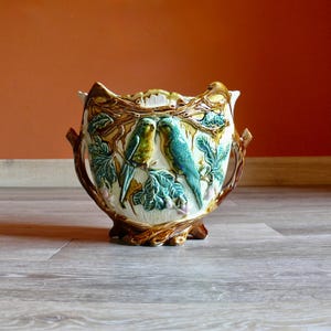 May include: A ceramic planter with a detailed bird and foliage design. The planter features two turquoise birds perched on a branch with green leaves. The rim and handles have a brown and gold finish.