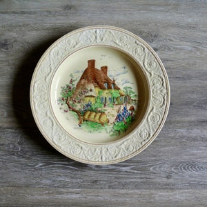 May include: A white ceramic plate with a raised rim and intricate floral design. The center of the plate features a colorful illustration of a cottage with a thatched roof, surrounded by flowers and greenery.