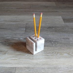 May include: A white marble pencil holder with a slot for a phone or other items. Two yellow pencils are in the holder.