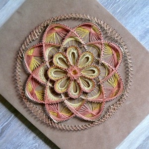 May include: A string art mandala with a flower design in shades of pink and yellow. The mandala is created with nails hammered into a piece of wood and string wrapped around the nails.