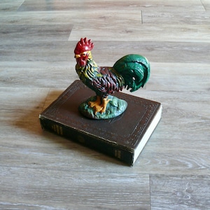 May include: A colorful rooster figurine made of metal, with a red comb and wattle, green tail feathers, and yellow feet. The rooster is perched on top of a brown leather-bound book.