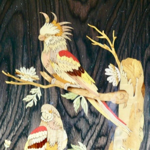 K&ouml;nnte beinhalten: Intricate Holzmarqueterie-Kunstwerk mit zwei V&ouml;geln, die auf &Auml;sten sitzen. Die V&ouml;gel sind aus verschiedenen Holzt&ouml;nen gefertigt, darunter Gelb, Rot und Braun. Der Hintergrund ist eine dunkle Holzmaserung.