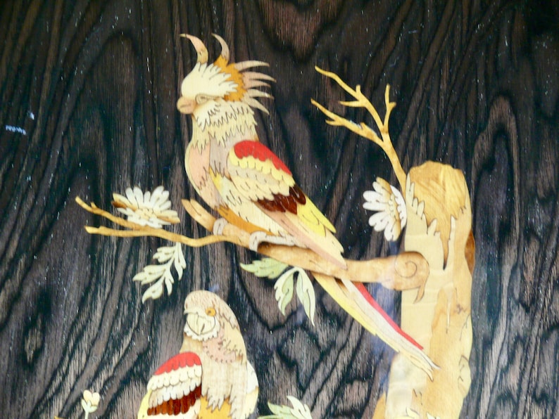 K&ouml;nnte beinhalten: Intricate Holzmarqueterie-Kunstwerk mit zwei V&ouml;geln, die auf &Auml;sten sitzen. Die V&ouml;gel sind aus verschiedenen Holzt&ouml;nen gefertigt, darunter Gelb, Rot und Braun. Der Hintergrund ist eine dunkle Holzmaserung.