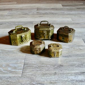 May include: Five antique brass hexagonal boxes of varying sizes. Each box has a hinged lid, a small handle, and a decorative latch. The boxes are arranged on a light-colored wooden surface, showcasing their intricate details and aged patina.