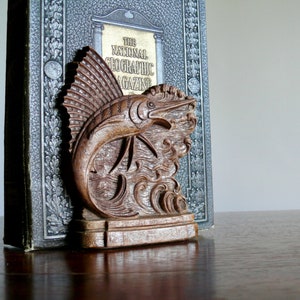 May include: A wooden bookend shaped like a marlin fish with detailed carvings. The bookend is resting on top of a book titled "The National Geographic Magazine".
