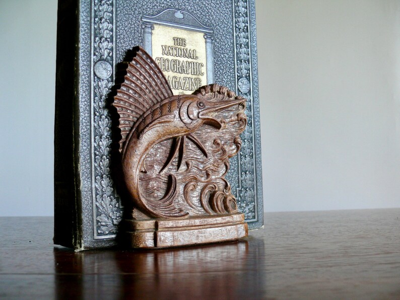 May include: A wooden bookend shaped like a marlin fish with detailed carvings. The bookend is resting on top of a book titled "The National Geographic Magazine".