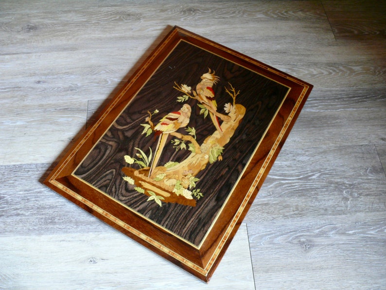 K&ouml;nnte beinhalten: Ein gerahmtes Holz-Marqueterie-Kunstwerk, das zwei V&ouml;gel auf einem Ast zeigt. Die V&ouml;gel sind aus verschiedenen Holzst&uuml;cken in verschiedenen Farben gefertigt, was einen Mosaik-Effekt erzeugt. Der Hintergrund ist eine dunkle Holzmaserung.