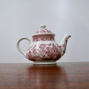 May include: A white and red ceramic teapot with a lid. The teapot features a detailed pattern of figures, buildings, and trees. The handle and spout are curved. The teapot is sitting on a wooden surface.