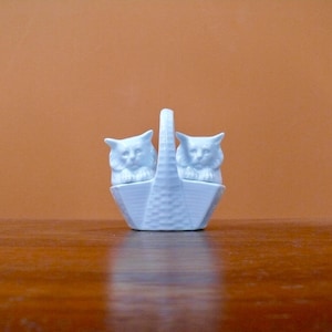 May include: A light blue ceramic basket-shaped container with a handle, holding two cat figurines. The cats have detailed facial features and are facing forward. The basket has a textured design. The background is a solid orange color.