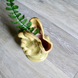 K&ouml;nnte beinhalten: Eine Vintage-Keramikvase in einem hellen Gelbgr&uuml;n, mit einem einzigartigen, gesichts&auml;hnlichen Design. Der Innenraum ist ein zartes Rosa. Ein Zweig gr&uuml;nes Kunstlaub befindet sich im Inneren der Vase. Die Vase steht auf einer hellen Holzoberfl&auml;che.