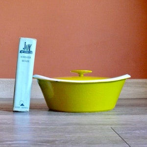 May include: A bright yellow ceramic casserole dish with a white lid and a small knob on top. The dish is sitting on a wooden surface. A cookbook titled "Joy of Cooking" by Rombauer and Becker is standing upright to the left of the dish.