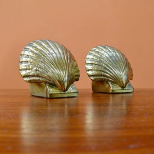 May include: Two brass-colored seashell bookends. Each bookend has a fan-shaped shell design with detailed ridges. The bookends are mounted on rectangular bases and sit on a wooden surface against a peach-colored background.