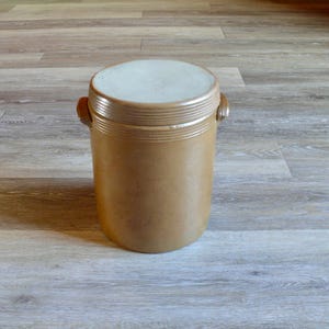 May include: A tan ceramic crock with a light-colored lid and two small handles. The cylindrical container has a ribbed design around the top and a smooth body. The crock is sitting on a wooden surface.