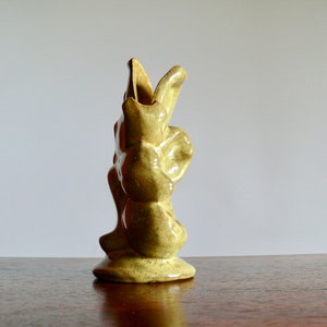 K&ouml;nnte beinhalten: Eine kleine, hellgr&uuml;ne Keramikvase mit einem einzigartigen, abstrakten Design. Die Vase hat eine strukturierte Oberfl&auml;che und eine ausgestellte Oberseite und steht auf einem runden Sockel. Die Vase ist etwa 15 cm hoch.