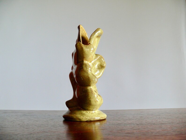 K&ouml;nnte beinhalten: Eine kleine, hellgr&uuml;ne Keramikvase mit einem einzigartigen, abstrakten Design. Die Vase hat eine strukturierte Oberfl&auml;che und eine ausgestellte Oberseite und steht auf einem runden Sockel. Die Vase ist etwa 15 cm hoch.