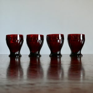 May include: Four ruby red vintage drinking glasses. Each glass has a unique, ribbed design on the body and a dark base. The glasses are arranged in a row on a wooden surface, reflecting the light.