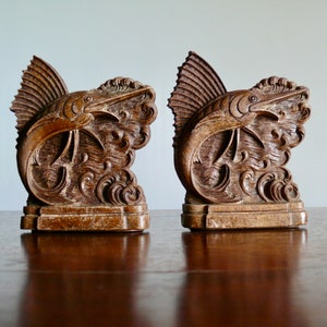 May include: A pair of wooden bookends featuring intricately carved marlin fish leaping out of the water. The bookends are a dark brown color and have a rustic, antique look.