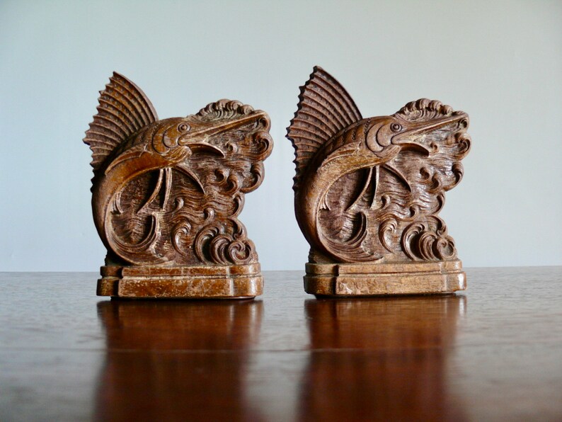 May include: A pair of wooden bookends featuring intricately carved marlin fish leaping out of the water. The bookends are a dark brown color and have a rustic, antique look.