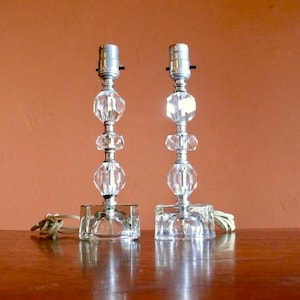 May include: Two clear glass table lamps with faceted crystal bases and silver metal accents. The lamps have a simple, elegant design.