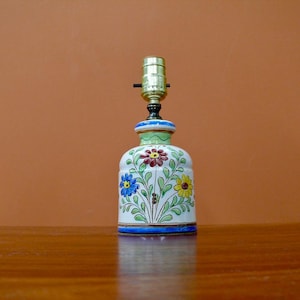 May include: A white ceramic table lamp with a green base and floral design. The lamp features blue, yellow, red, and green flowers with green leaves. The lamp has a gold trim around the base and a gold metal lampshade.