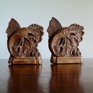 May include: A pair of wooden bookends shaped like marlin fish leaping out of the water. The bookends are a dark brown color and have intricate details carved into the wood.