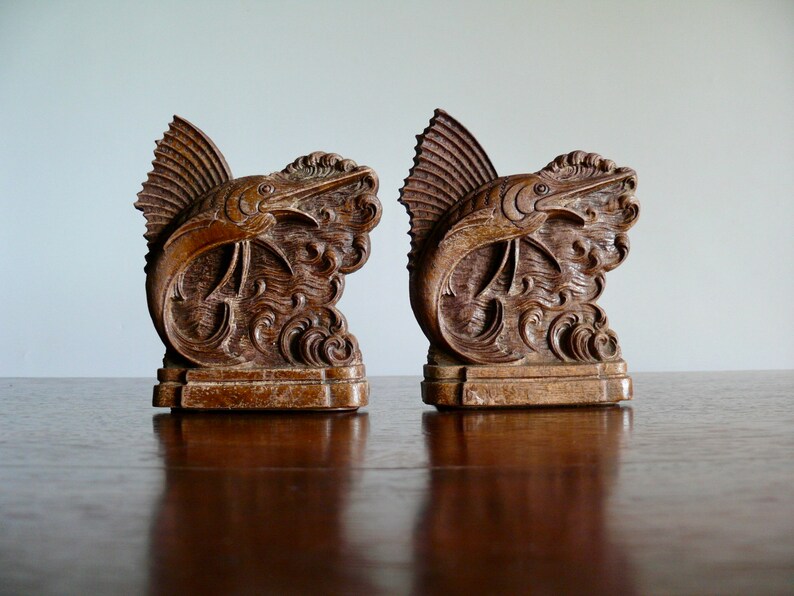May include: A pair of wooden bookends shaped like marlin fish leaping out of the water. The bookends are a dark brown color and have intricate details carved into the wood.