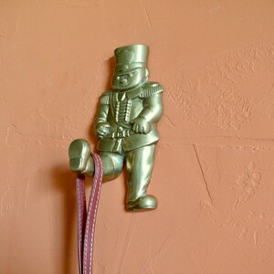 May include: A gold-toned metal wall hook shaped like a toy soldier. The soldier is wearing a hat and uniform and is holding a rifle. The hook is attached to a wall with a brown textured surface.