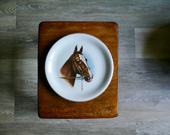 May include: A white ceramic plate with a brown and white horse head illustration. The plate is on a wooden surface.