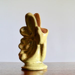 K&ouml;nnte beinhalten: Eine Keramikvase in Form einer Hand, die ein Friedenszeichen zeigt. Die Vase hat eine hellgelb-gr&uuml;ne Farbe mit gl&auml;nzendem Finish. Das Innere ist ein dunkleres, r&ouml;tlich-braunes. Die Vase hat einen runden Sockel und ein einzigartiges, k&uuml;nstlerisches Design.