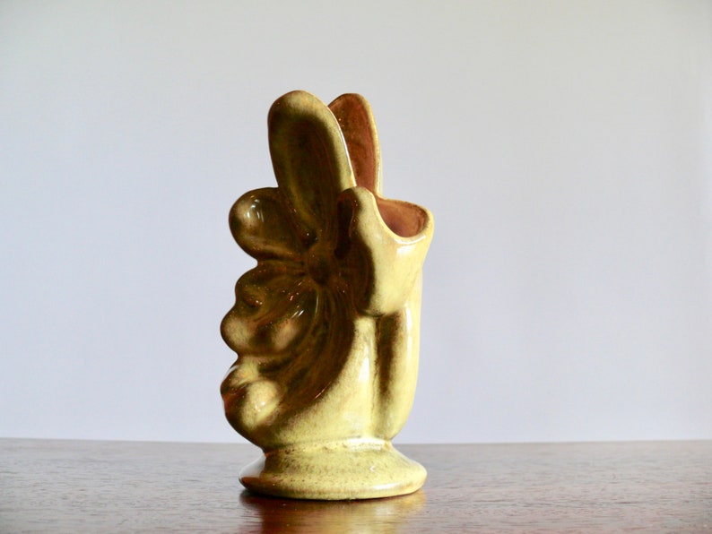 K&ouml;nnte beinhalten: Eine Keramikvase in Form einer Hand, die ein Friedenszeichen zeigt. Die Vase hat eine hellgelb-gr&uuml;ne Farbe mit gl&auml;nzendem Finish. Das Innere ist ein dunkleres, r&ouml;tlich-braunes. Die Vase hat einen runden Sockel und ein einzigartiges, k&uuml;nstlerisches Design.