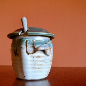 May include: A ceramic honey pot with a lid and a wooden dipper. The pot is decorated with a green and brown glaze and has a white background.