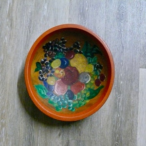 May include: A wooden bowl with a painted design of fruit, including grapes, apples, and pears. The bowl is orange with a brown rim.