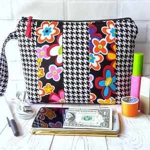 Puede incluir: Una bolsa con cremallera de tela de pata de gallo en blanco y negro con un estampado floral en la parte delantera. La bolsa tiene un tirador de cremallera rojo y un asa negra. La bolsa está sobre una superficie blanca con un teléfono móvil, un bolígrafo, un billete de dólar y otros artículos.