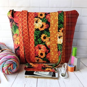 May include: A large tote bag with a patchwork design featuring fall-themed patterns. The bag includes panels with pumpkins, sunflowers, and plaid. The bag has orange straps and a matching wristlet. A variety of accessories are next to the bag.