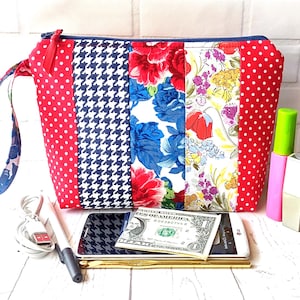 Puede incluir: Una bolsa de cremallera roja, blanca y azul con un asa. La bolsa está llena de varios artículos, incluyendo un teléfono, un cargador, un bolígrafo y un billete de un dólar.