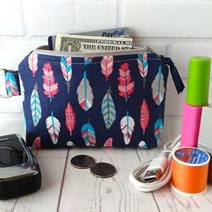 Könnte beinhalten: Eine marineblaue Reißverschlusstasche mit einem Feder-Muster in Rosa, Weiß und Blau. Die Tasche ist offen und zeigt einen Dollarschein und eine Kreditkarte. Ein Nokia-Telefon, Münzen und anderes Zubehör liegen neben der Tasche.