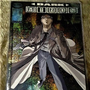 Puede incluir: Una portada de cómic con acabado holográfico que presenta a un hombre con un abrigo largo de pie en un entorno urbano. El título del cómic es "Dark Minds" y la portada es de Image Comics.