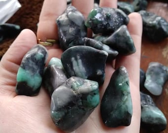 May include: A handful of raw emerald stones, showcasing a mix of dark green and gray hues. The rough-cut gemstones have a natural, unpolished appearance, with visible textures and variations in color. The emeralds are displayed against a neutral background.