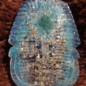 Puede incluir: Un colgante de resina azul transparente con forma de sarcófago egipcio con purpurina dorada incrustada en su interior.